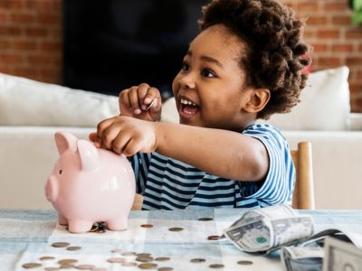 Child joyfully saving money in a pink piggy bank surrounded by coins and dollar bills, illustrating the financial benefits of water filtration systems.