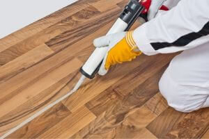 A person in yellow gloves uses a caulk gun to seal a seam on a wooden floor