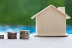 Stacks of coins next to a small model house, symbolizing rising utility costs related to HVAC inefficiency and ductwork issues.