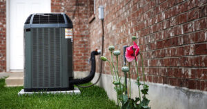 Air conditioner unit beside flowering poppy plants and textured brick wall in a green grass outdoor setting.
