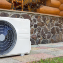 Air conditioning unit against a stone wall of a log cabin, showcasing modern HVAC technology in a rustic setting.