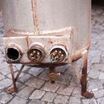 Rusty metallic tank on cobblestone with corrosion and plumbing connections. PAC Plumbing maintenance services focus.