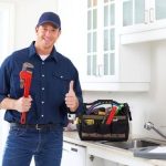 Male plumber with adjustable wrench and toolbox, giving a thumbs-up near a kitchen sink for PAC Plumbing services.