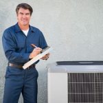 Male HVAC technician in blue uniform stands by air conditioning unit, clipboard in hand, representing PAC Plumbing services.