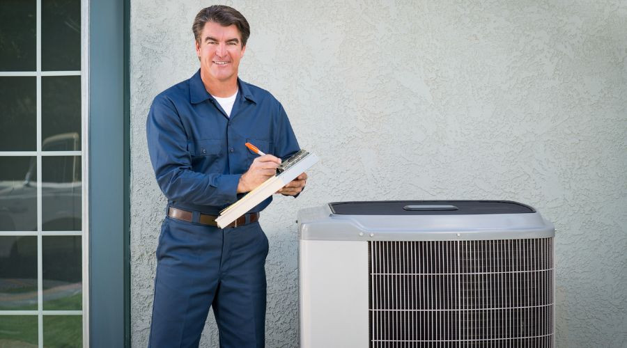 Technician in blue uniform next to gray air conditioning unit for PAC Plumbing, Heating, Air Conditioning services.