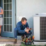 Technician servicing an air conditioning unit, with toolbox and homeowner observing in a practical maintenance setting.