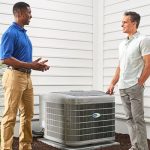 Two men discussing HVAC services near an air conditioning unit, with a home exterior in the background.