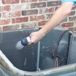 Person using a handheld device to inspect an air conditioner unit against a brick wall, showcasing PAC Plumbing services.