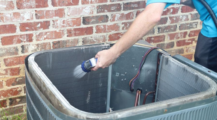 Person cleaning an air conditioning unit, showcasing internal components and water spraying action against a brick wall.