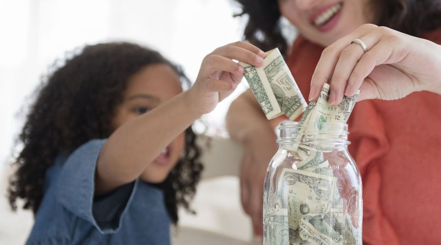 An adult woman guides a child inserting dollar bills into a glass jar, promoting financial education and family bonding.