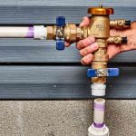 Brass plumbing valve with colorful plastic handles, held in hand against PVC piping and connectors for maintenance tasks.